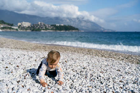 Kid crawls along the pebble beach against the background of the sea and mountainsの写真素材