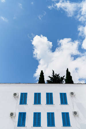 White two-story building with closed blue shutters on the windows against a background of blue sky and white cloudsの写真素材
