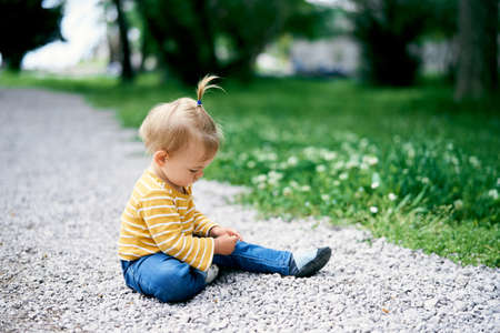 Little girl with a ponytail on her head sits on a gravel path in a green park
