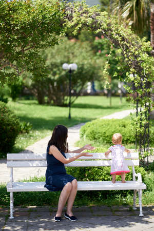 Little girl stands on a park bench next to her mother sittingの写真素材