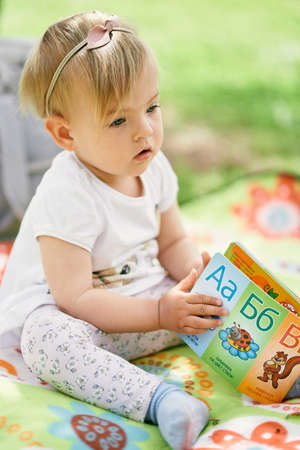 Little girl sits on a colored blanket on a green lawn and holds a childrens book in her handsの写真素材