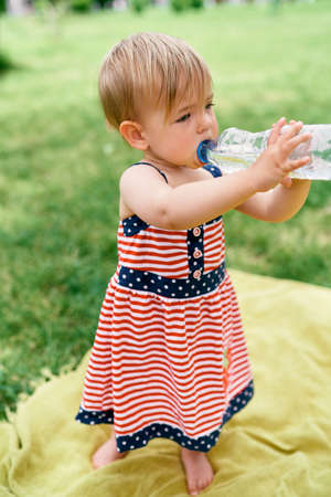 Little girl drinks water from a bottle standing on a blanket on the lawnの写真素材