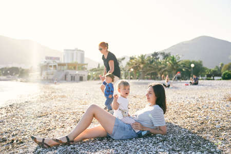 Mom lies on a pebble beach and holds a little girl on her stomach near the seaの写真素材
