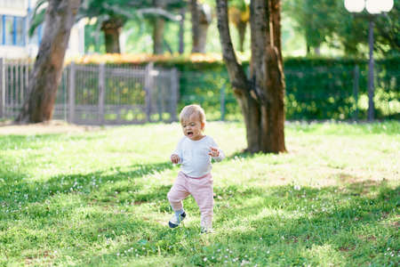 Sad kid walks on a green lawn with wildflowers against the backdrop of a house and a fenceの写真素材