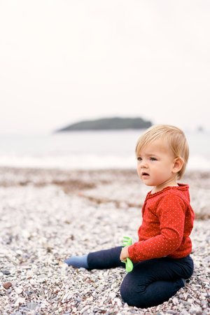 Small child sits on a pebble beach and holds a toy rake in his handの写真素材