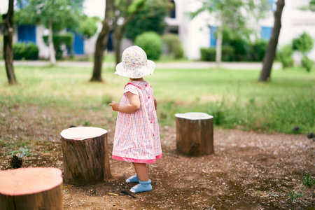 Little girl in a hat and dress stands in the yard near the stumps, turning her headの写真素材