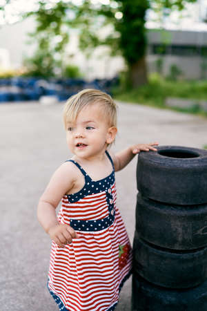 Little girl in a dress stands near a pile of small tires in a car parkingの写真素材