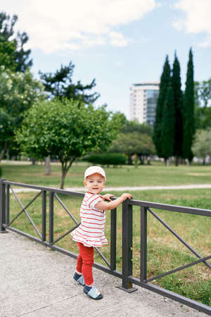 Little girl in a panama hat stands near a metal fence in a green parkの写真素材