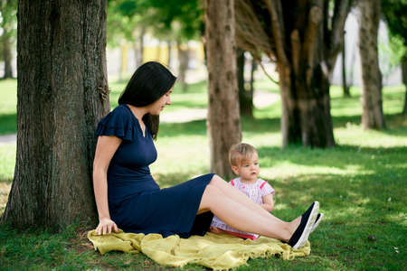 Mom with a little girl are sitting on a blanket on a green lawn under a treeの写真素材