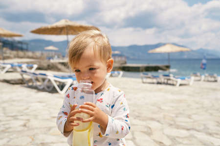 Kid holds a bottle of water while standing on a cobbled beach with sun loungersの写真素材