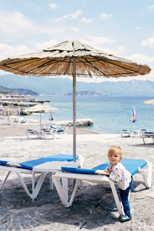 Kid stands near a sun lounger under a straw umbrella on the beach near the waterの写真素材