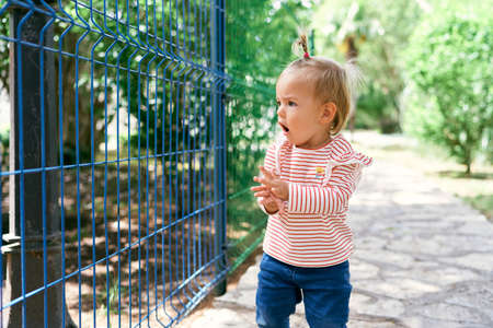 Little girl stands on a paved path near a metal fence with an open mouthの写真素材