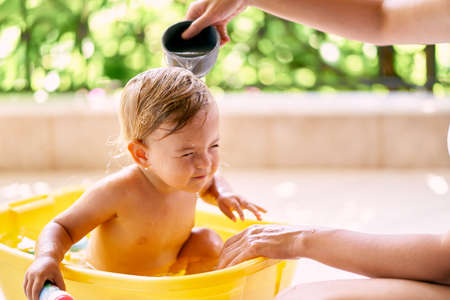 Mom pours water from a ladle to a closed-eyed child sitting in a bowlの写真素材