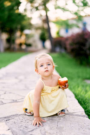 Little girl with an apple in her hand squatted on a paved pathの写真素材