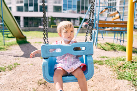 Little girl sits on a swing on a playground near an apartment buildingの写真素材