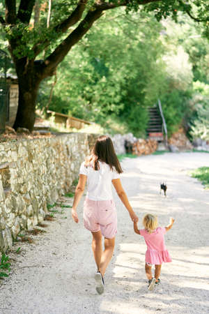 Mom leads a little girl by the hand along the gravel path in the parkの写真素材