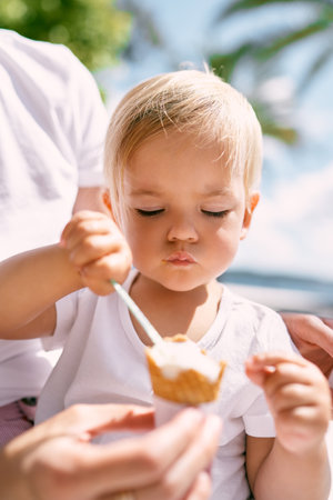 Little girl carefully takes ice cream with a spoon, sitting on mom lapの写真素材