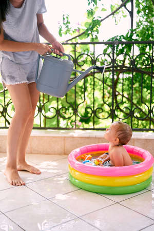 Mom with a watering can stands over a little girl in an inflatable pool on the balcony