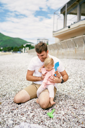 Dad holds little girl on his lap while sitting on pebble beachの写真素材