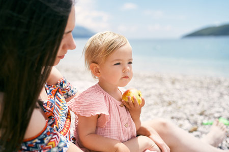 Little girl with an apple in her hand sits on the beach next to her motherの写真素材