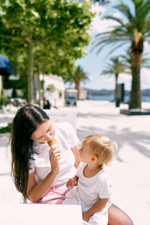Mom tastes ice cream in front of a little girl sitting on her lap in a cafe on the beach