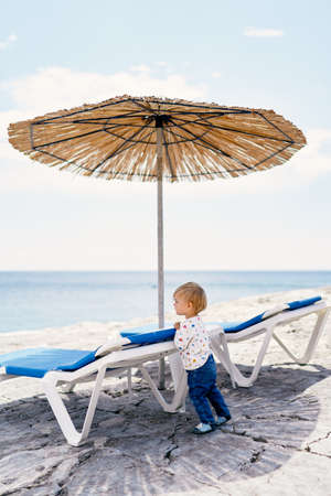 Kid stands leaning on a sun lounger under a thatched beach umbrella and looks at the seaの写真素材