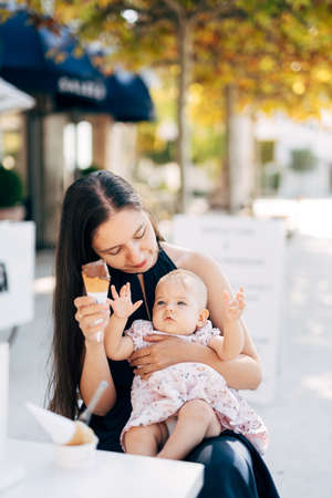 Mom with a little girl on her knees holds ice cream in her hand while sitting in a cafeの写真素材