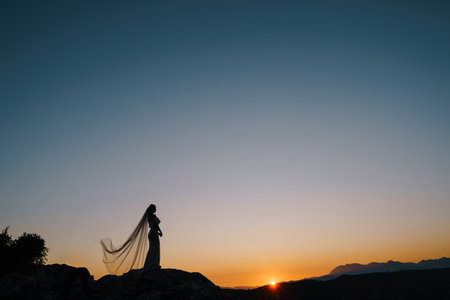 Silhouettes of a bride in a fluttering veil standing on a mountain and looking at the sunsetの写真素材