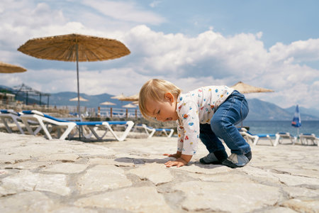 Kid is on all fours on a cobbled beach with sun loungers and umbrellasの写真素材