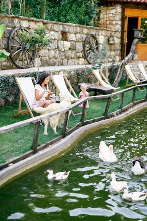Mom with a little girl sit on a green lawn by a pond with ducksの写真素材