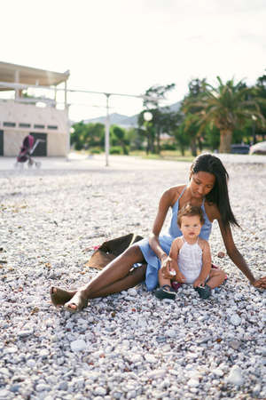 Mom sits on the beach and holds out a pebble to a little girlの写真素材