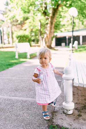 Little girl with a bottle of water in her hands stands by a park benchの写真素材