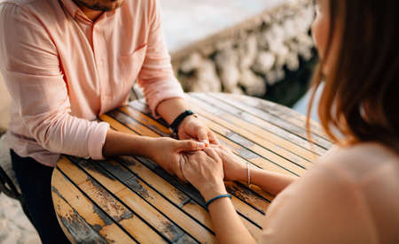Man in a pastel shirt and a wristwatch holds the girls hands with bracelets behind a wooden table....
