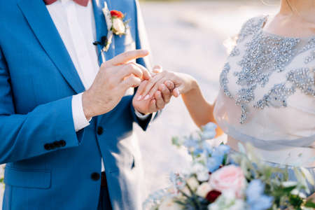 Groom in a blue suit puts a wedding ring on the brides finger with a bouquet of flowers. Close-upの写真素材