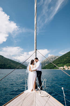 The bride and groom are hugging while standing on the bow of a white yacht sailing in the Bay of...