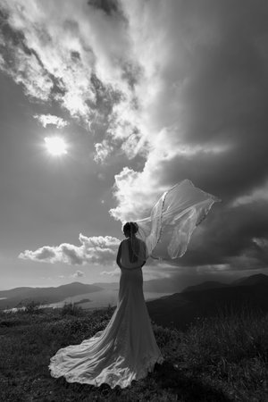 Bride in a veil fluttering in the wind and a long white dress stands on a mountain against the background of the sea. Black and white photo. Back viewの写真素材