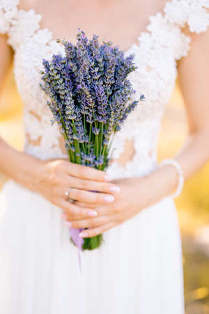 Bride in a white lace dress holds a lavender bouquet tied with a lilac ribbon. Close-upの写真素材