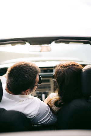 The bride and the groom are sitting in an open car on the picturesque road in the mountains in...