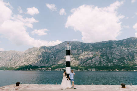 The bride and groom are standing and holding hands on the pier near the lighthouse in the Bay of Kotorの写真素材