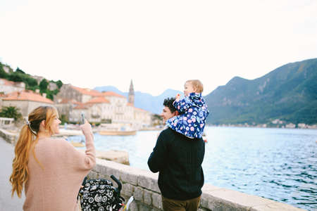 Mom drives the stroller, dad carries the daughter on his shoulders. Family walks along the coast against the backdrop of the sea, mountains and houses of the town of Perastの写真素材