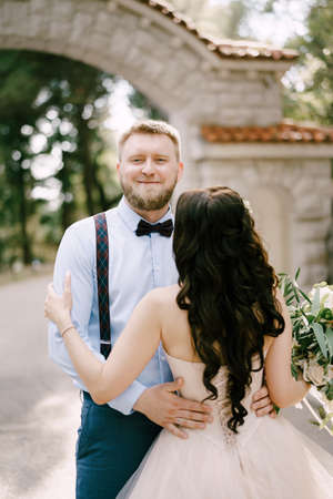 The bride hugs the groom in the park next to a beautiful brick arch, back viewの写真素材
