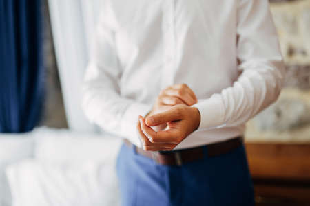 Man in blue trousers buttons a button on the cuff of a white shirt. Close up