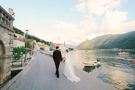 Bride and groom walk along the pier holding hands, against the backdrop of the bay, boats and ancient buildingsの写真素材