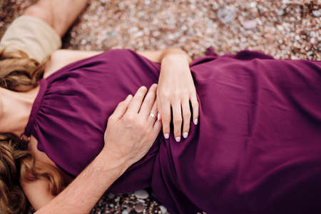 Bride in a burgundy dress lies on a pebble with her hands folded on her stomach. Close-up