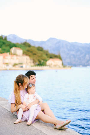 Dad, mom and little daughter are sitting on the pier by the sea against the backdrop of the city of Perast and the mountainsの写真素材