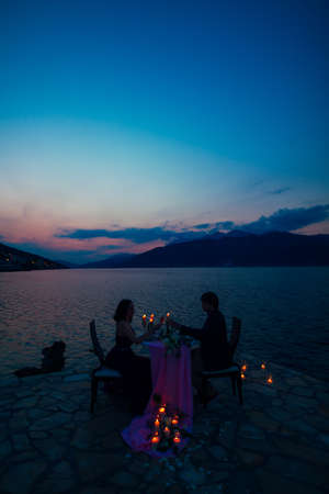 Man and woman sit at a table and clink glasses on the pier against the background of the sea and mountains at night by burning candlesの写真素材