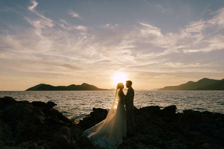 Groom and bride stand on the rocky coast by the sea at sunsetの写真素材