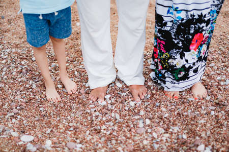 Bare feet of a man in light trousers, a woman in a colorful skirt and a child in shorts stand on a pebble beach. Close-upの写真素材