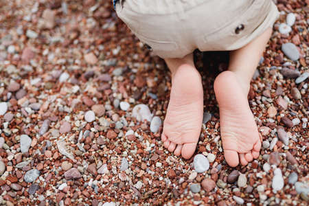 Child in short light shorts sits on his knees on a pebbled beach. Close-upの写真素材