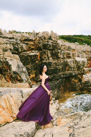 Girl in a beautiful lilac dress fluttering in the wind stands on a rock against the background of seething waterの写真素材